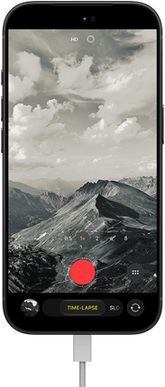 iPhone 18 Pro mostrato di fronte, sul display un paesaggio montuoso, con fotocamera in modalità Time-Lapse e cavo di alimentazione collegato alla porta di ricarica